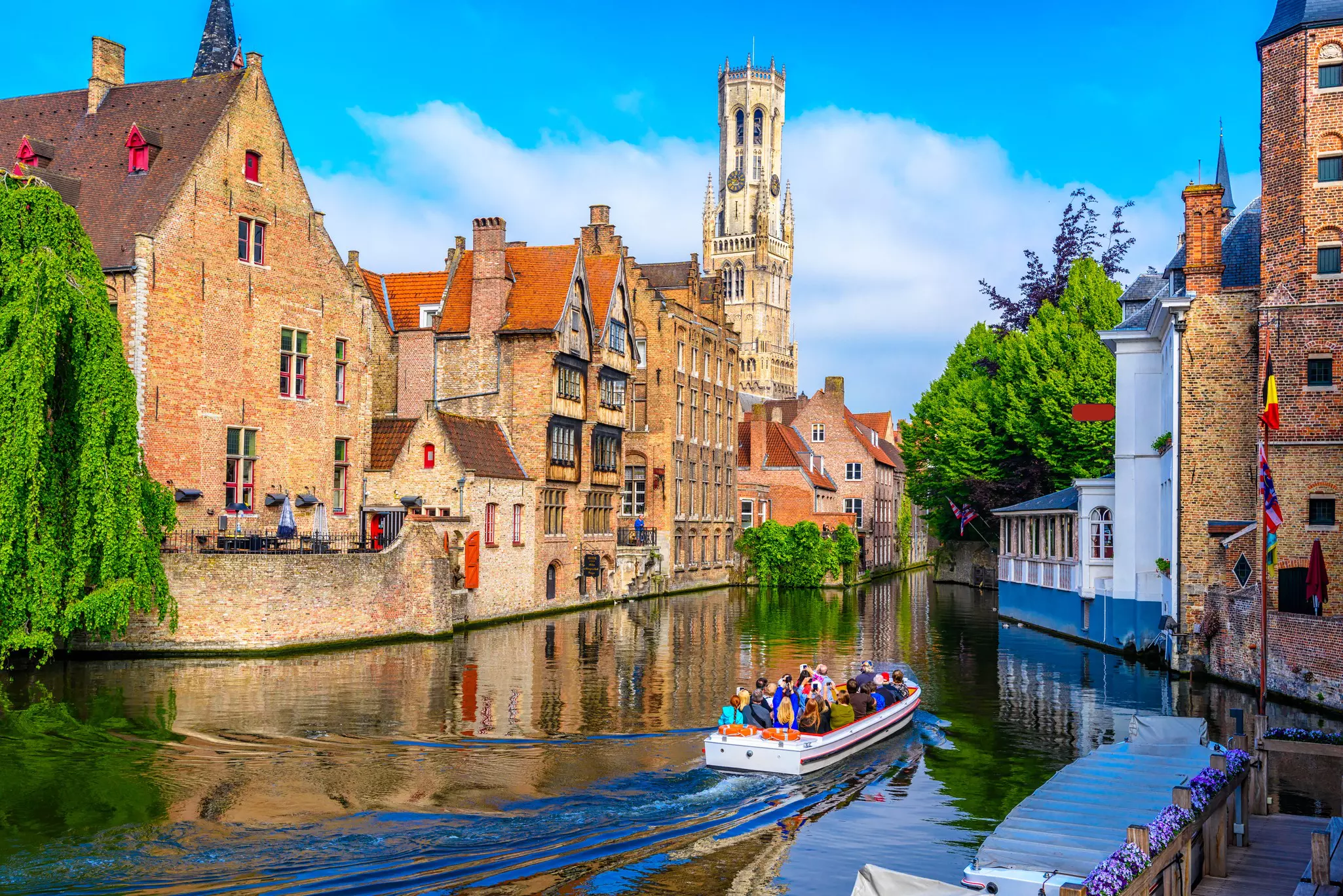 A small boat loaded with tourists travels along a canal in a medieval city with a large tower overlooking it