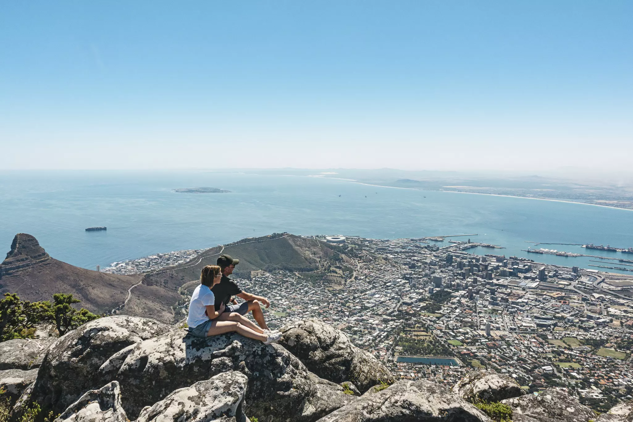 A couple sit on a rock overlooking a sprawling city below
