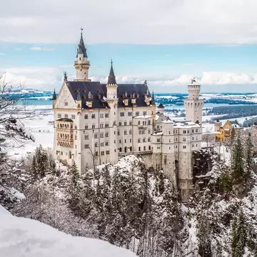 A white castle with pointed towers stands on a forested ridge covered in snow, with a flat wintry plain and lakes behind.