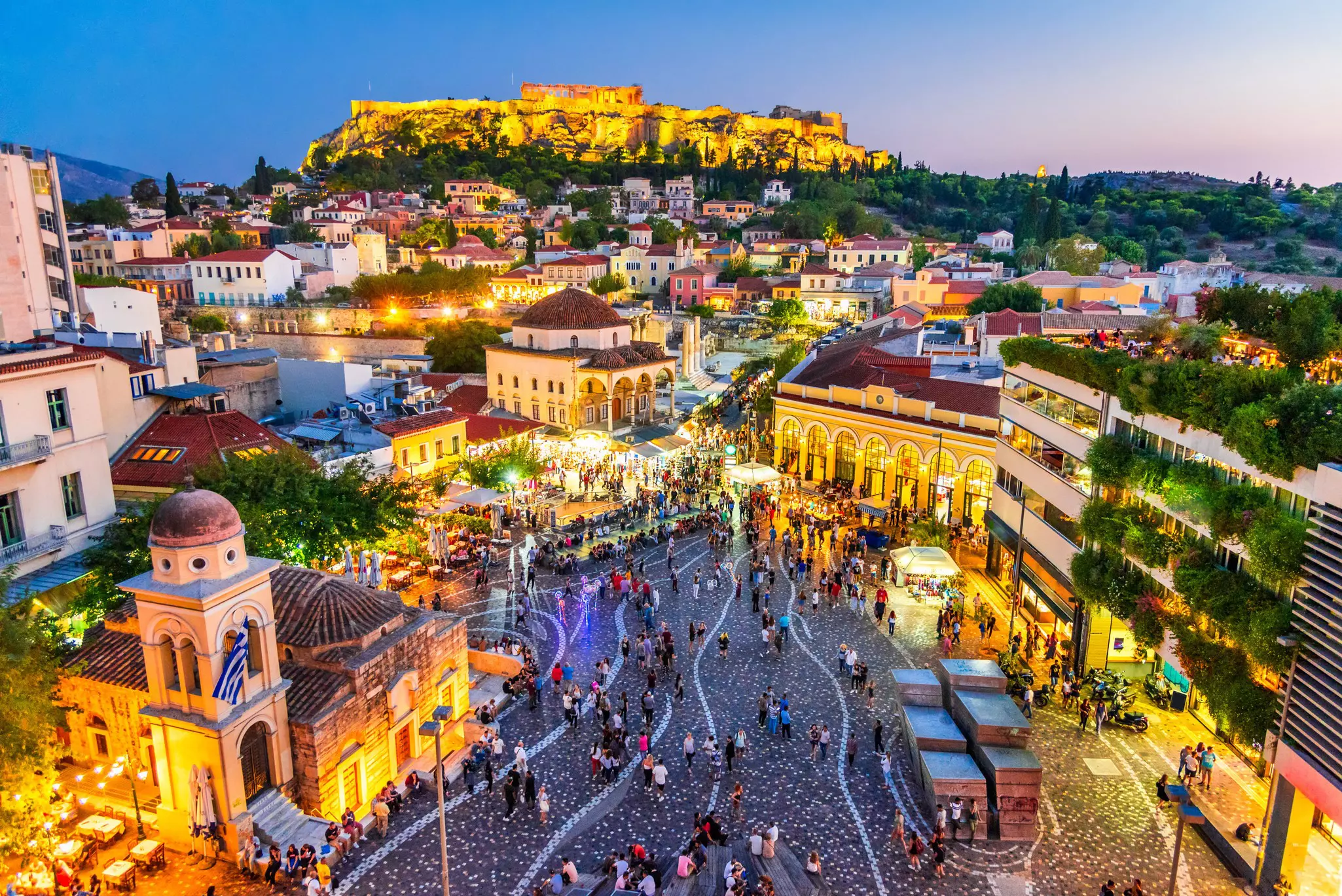 People mill through a city square in the evening, shopping at the market or dining at nearby restaurants. An ancient structure stands on a hill above.