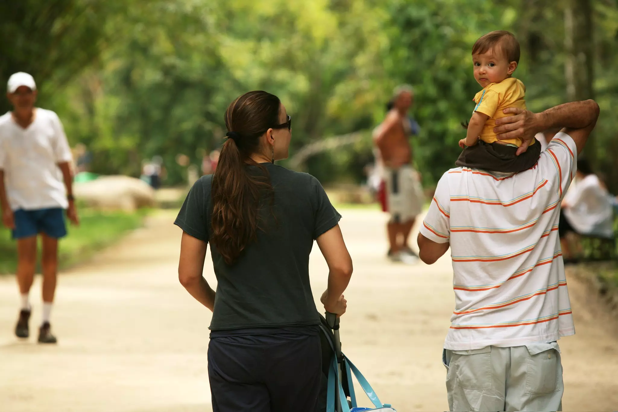 Two parents are seen from behind walking down a path in a city park. A toddler sits on one of their shoulders, and turns to face the camera.