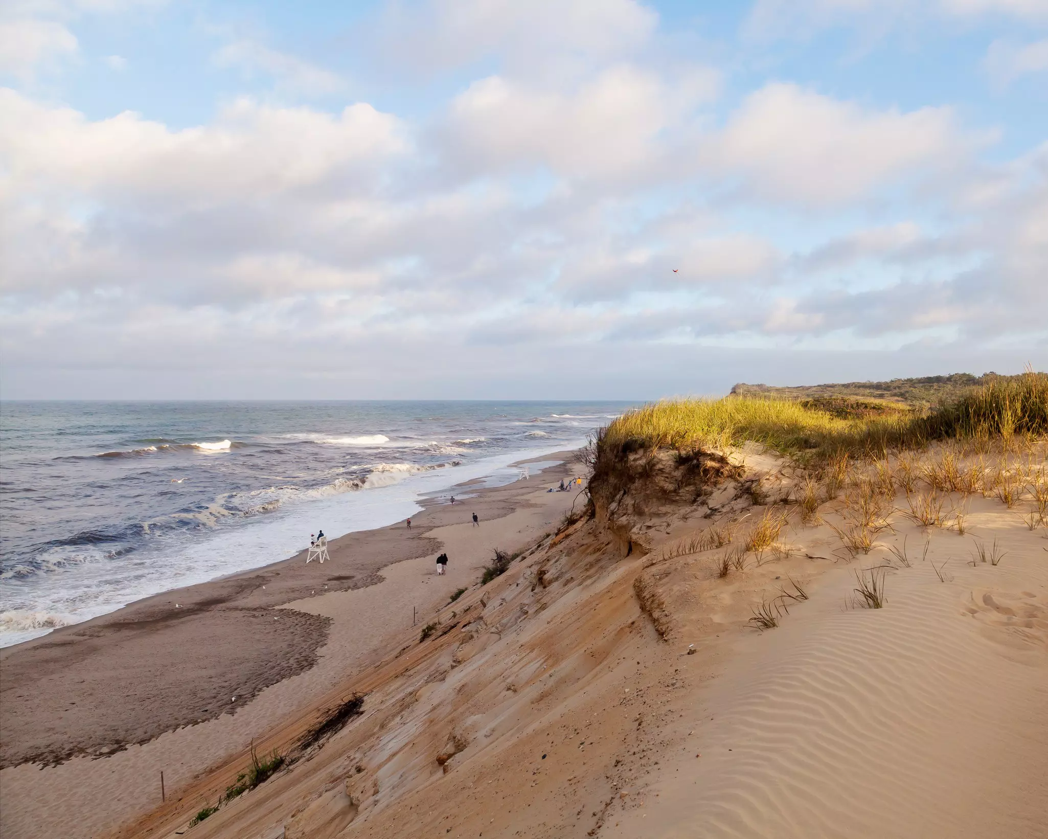 A wide view of dunes at sunset, rising over a beach lapped by whitecapped waves.