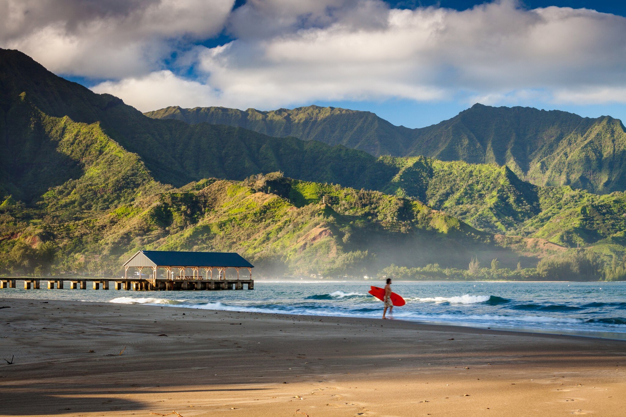 A person holds a surfboard on an empty beach. Lush cliffs are seen across the water.