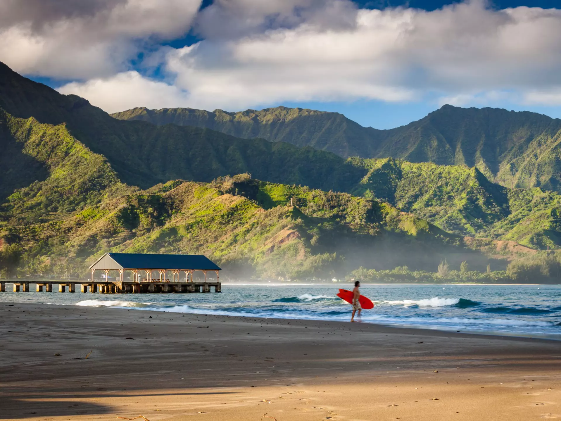 A person holds a surfboard on an empty beach. Lush cliffs are seen across the water.
