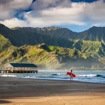 A person holds a surfboard on an empty beach. Lush cliffs are seen across the water.
