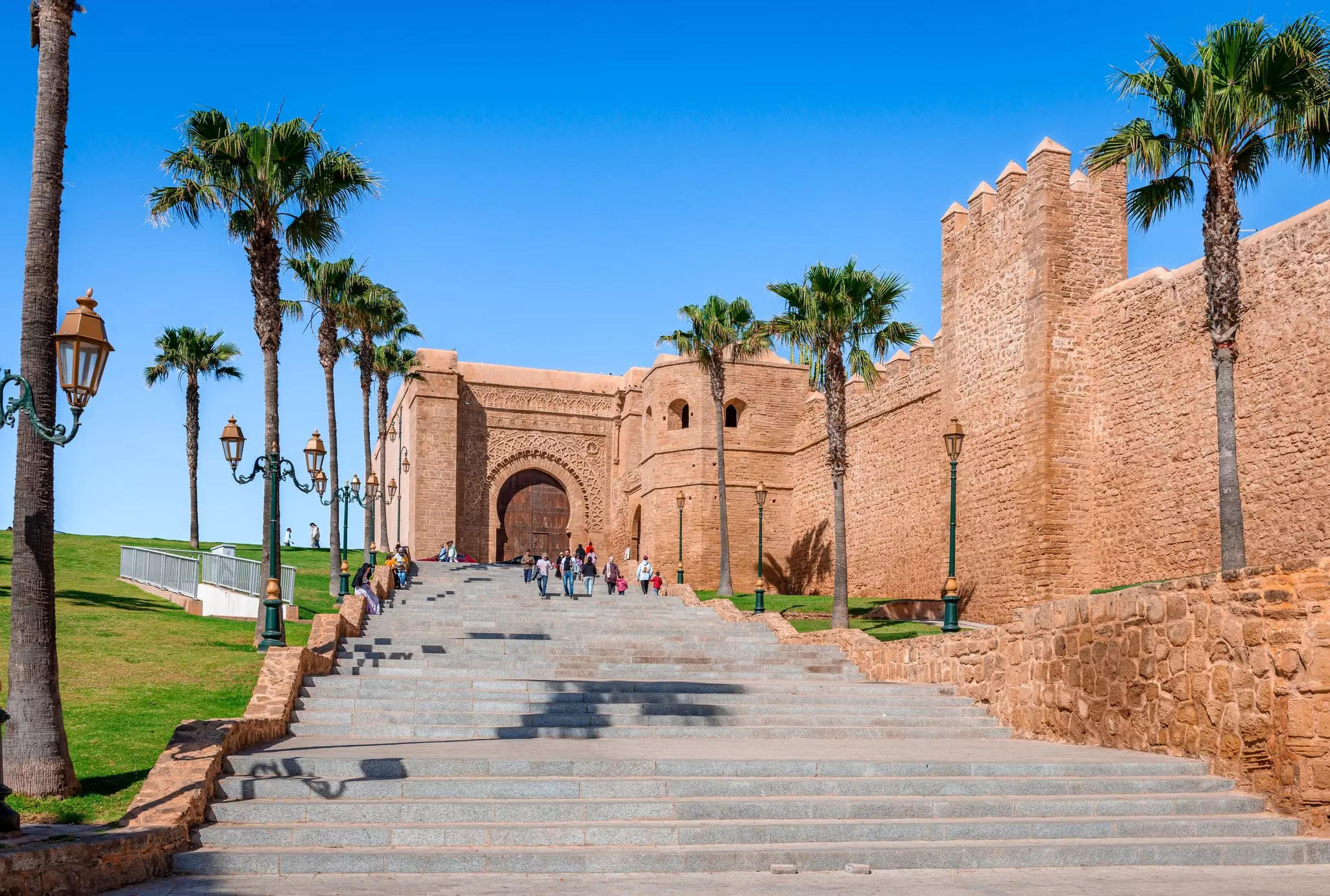 People walking up numerous stairs leading to the large doors of a brick building. Palm trees are on both sides of the stairway