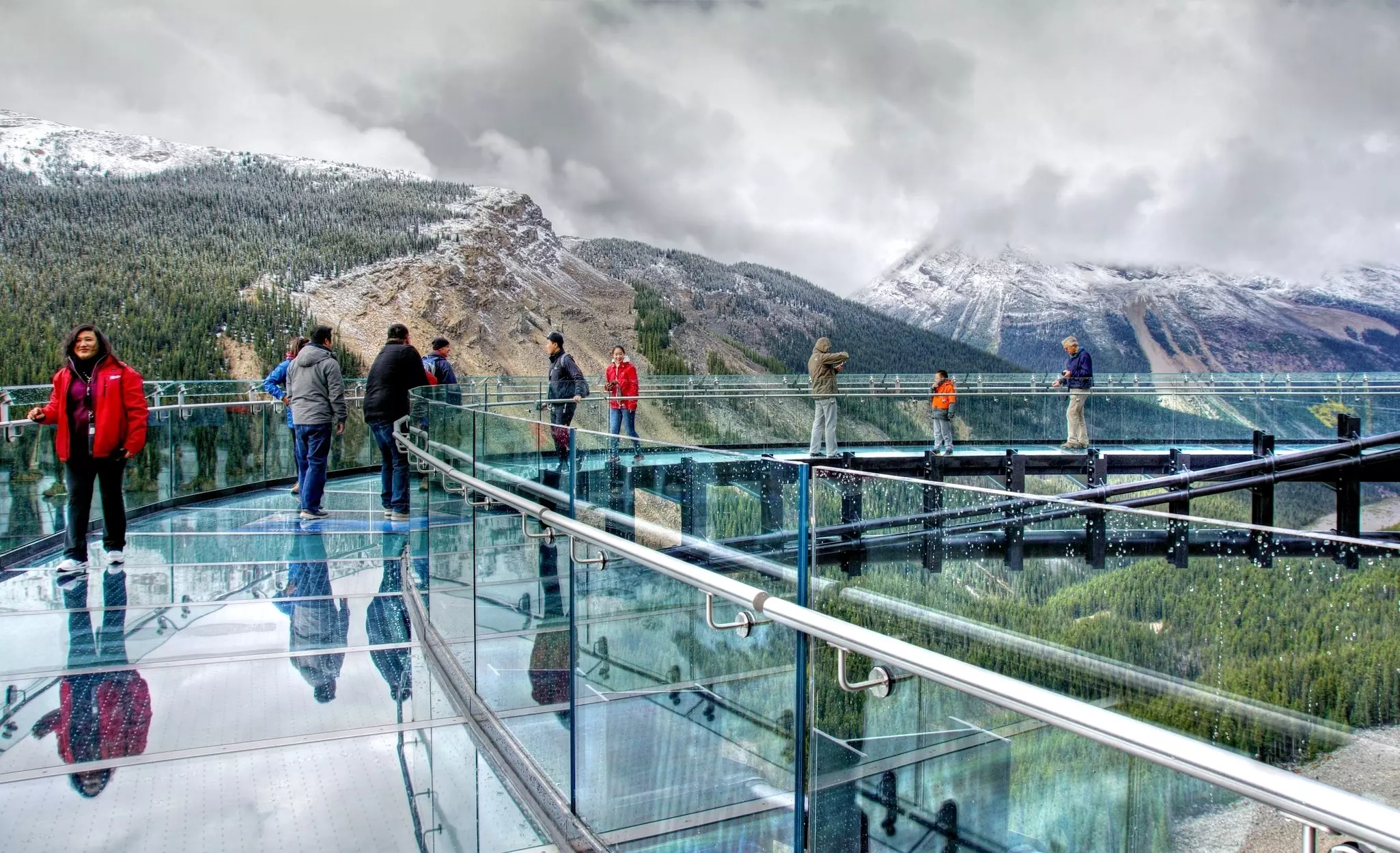People are seen on a translucent platform hanging over a mountain valley with snowy peaks in the distance.