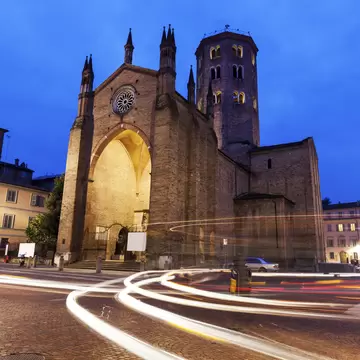 Brick Gothic basilica with an octagonal bell tower, lit at dusk, with streaks of car headlights on a cobbled street.
