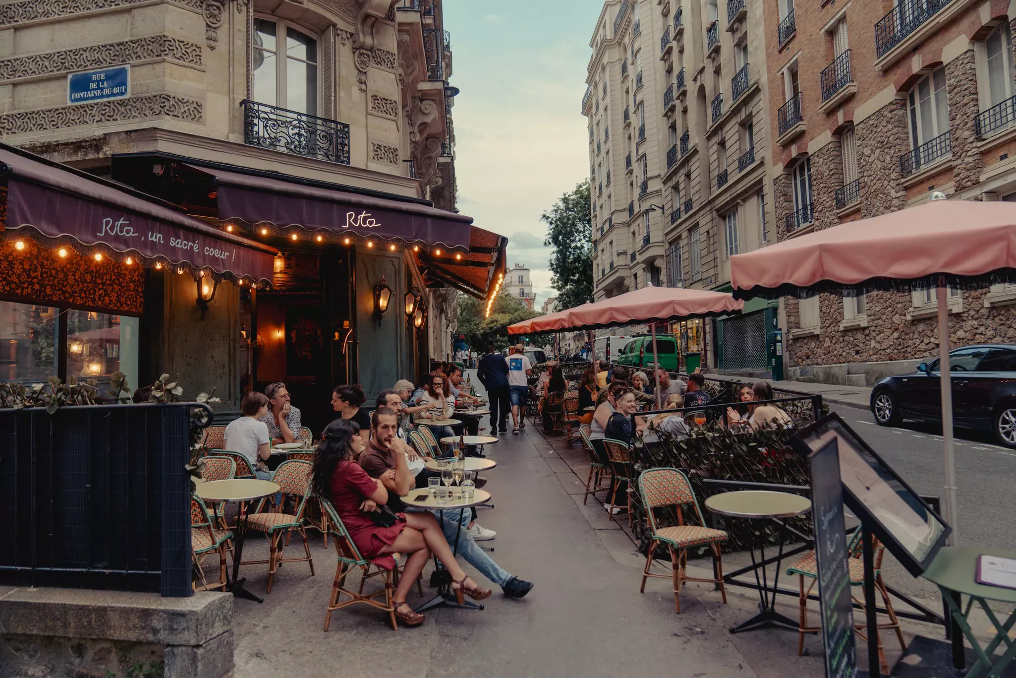 People sitting at a sidewalk cafe in Paris in early evening; there are lights around the awning of the cafe.