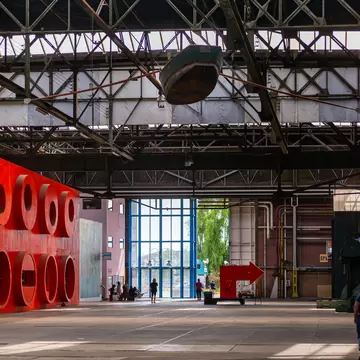 Cavernous steel-roofed warehouse at NDSM Wharf, Amsterdam, with red sculpture panels and a few people near a glass doorway.