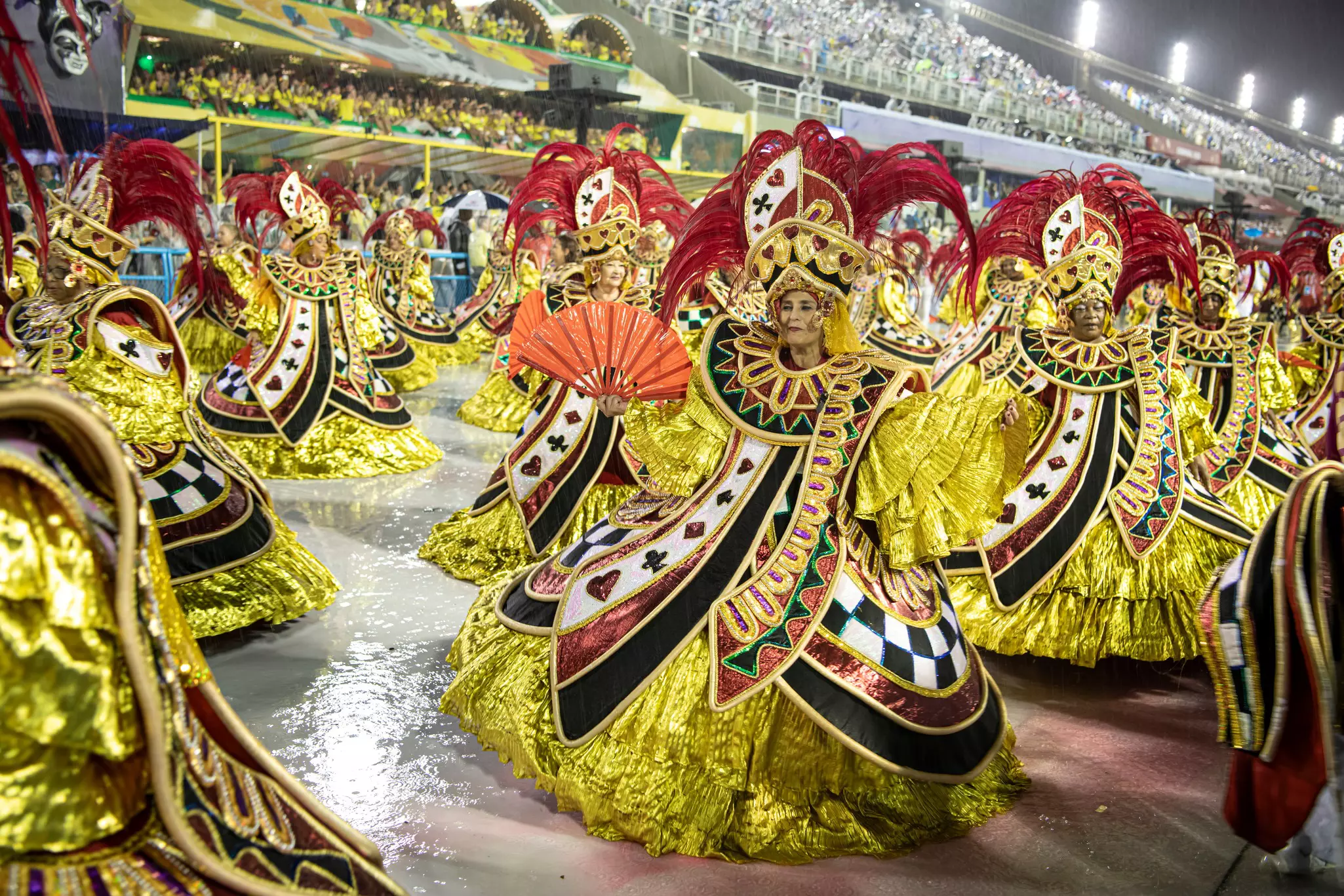 Dancers in elaborate gold costumes and feathered headdresses participate in parade in a stadium, as thousands of spectators look on.