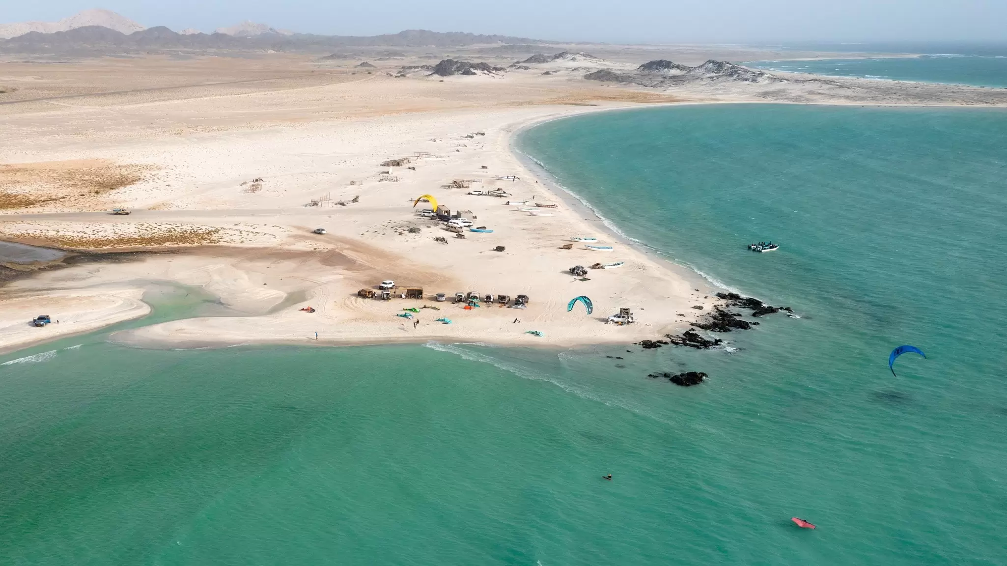 Aerial view of a sandy spit and turquoise lagoon with kitesurfers on the water off a desert island coast.