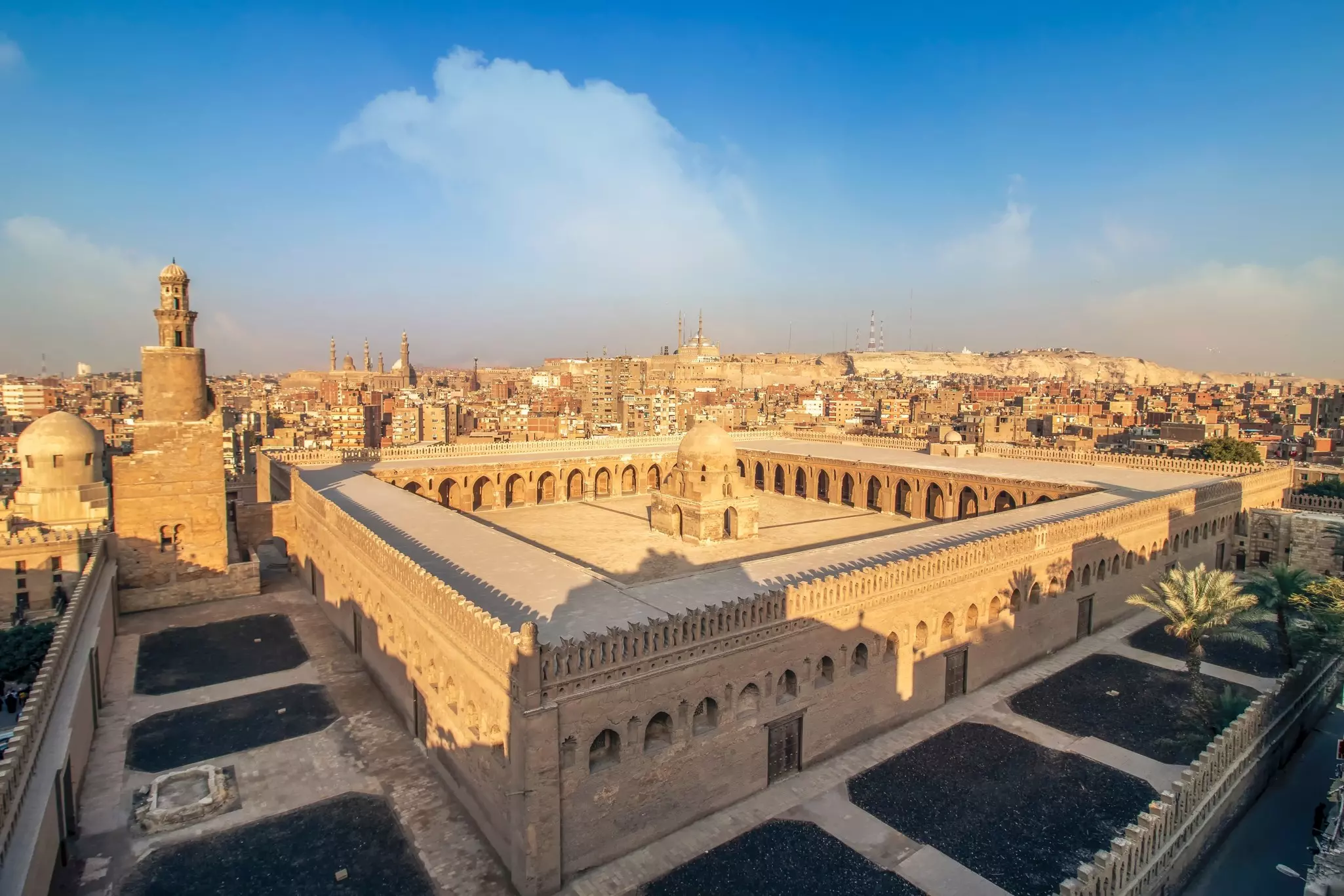 A large square building with a courtyard is in the foreground; the skyline of Cairo, Egypt, is visible beyond its walls.