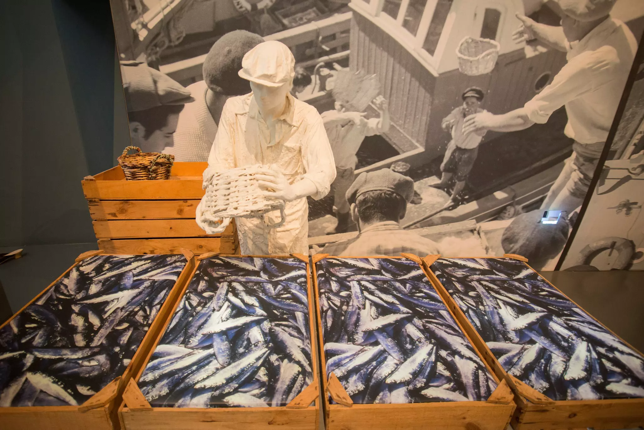 A white figure of a fish canning factory worker in a museum exhibit. Four crates of fake fish are in front of him.