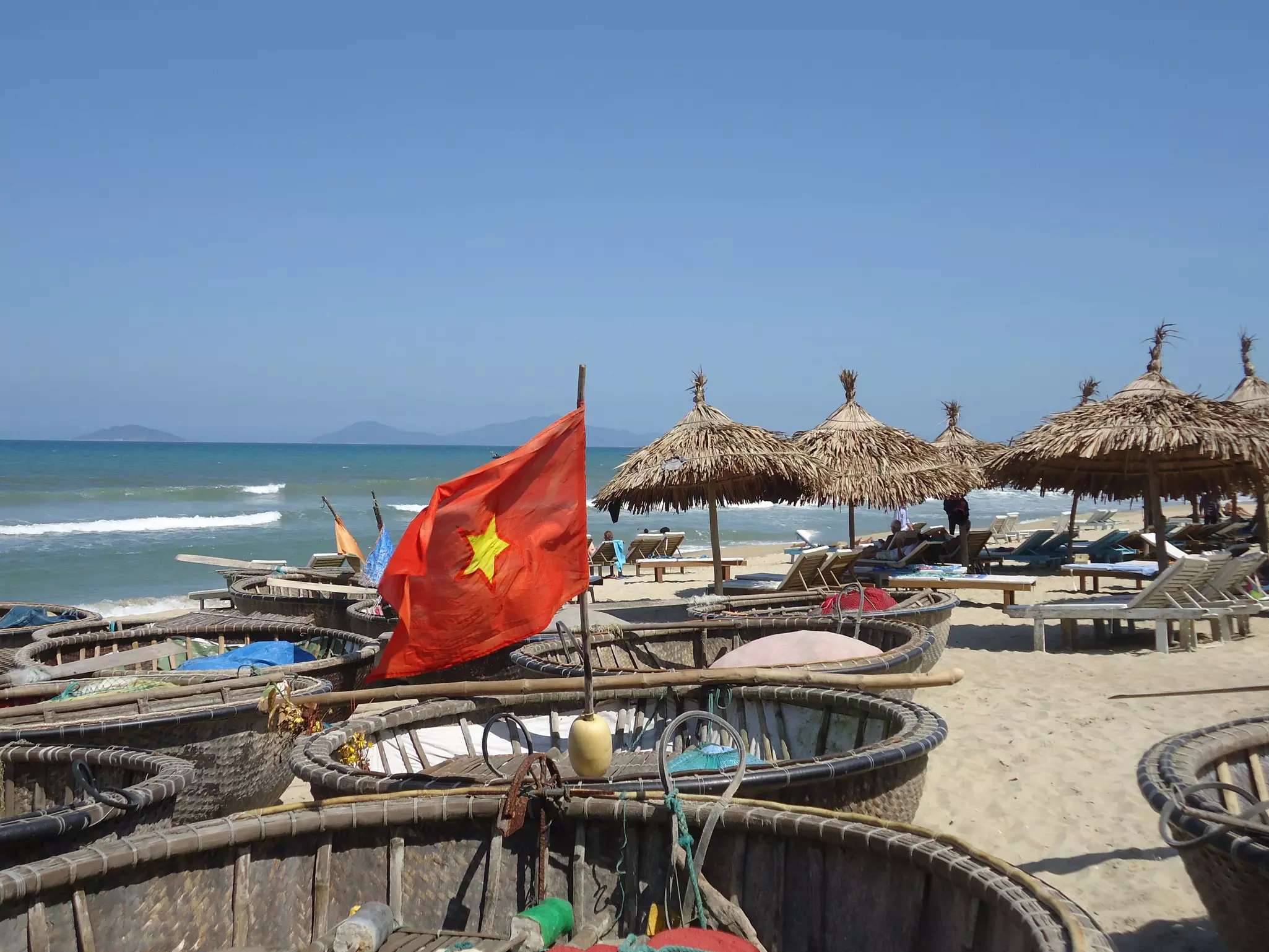 A Vietnamese flag flies above a basket boat on the beach, with some sunshades and sunloungers in the background
