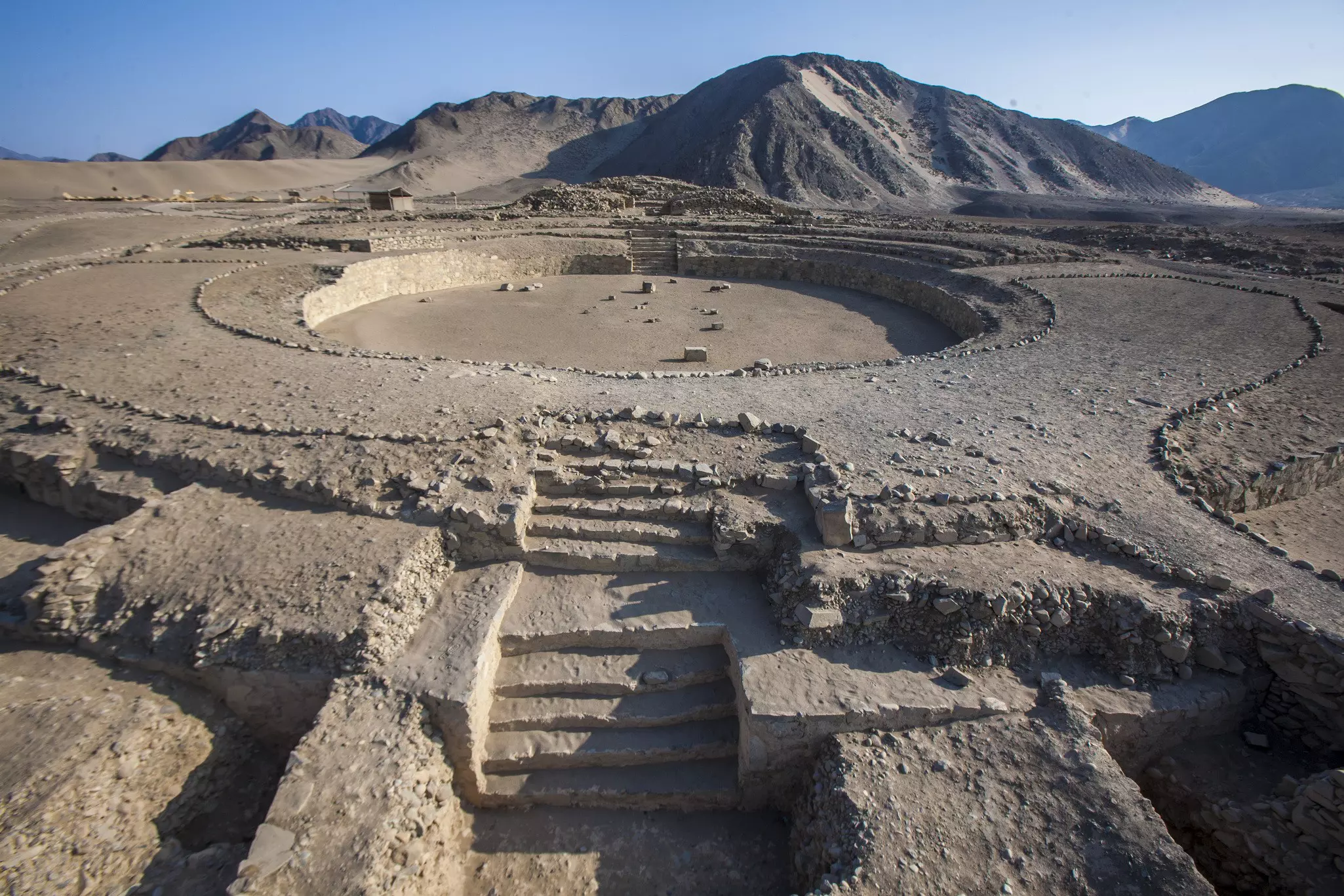 Steps lead to a massive circular opening at an ancient archeological site in the desert, with dry hills in the background
