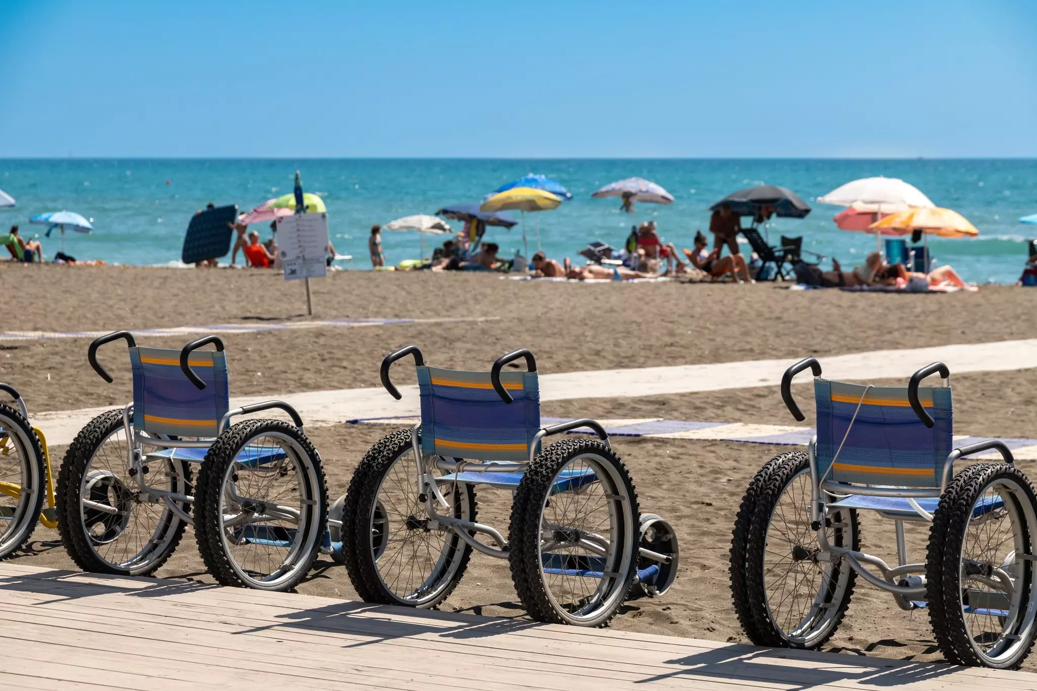 A row of four beach-accessible wheelchairs with fat tires standing at the promenade end of a beach