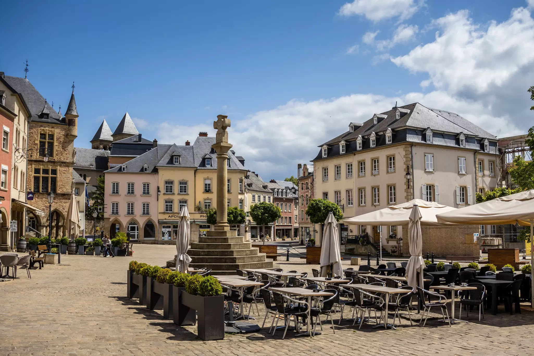 A small market square surrounded by old buildings. Restaurant tables are set up in the center of the square.