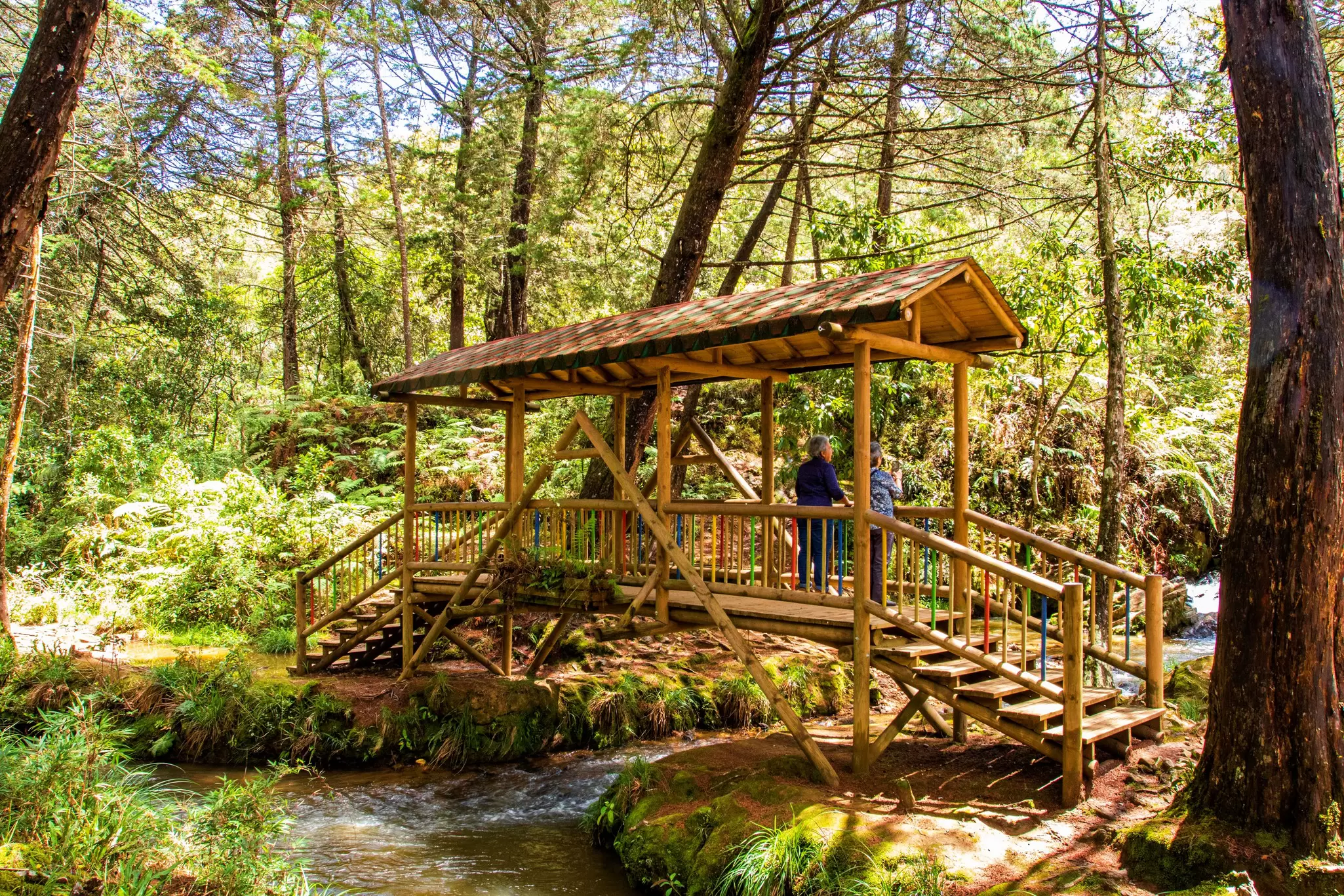 Two tourists standing on a sheltered wooden bridge above a flowing river in Parque Arvi in Santa Elena, Colombia.