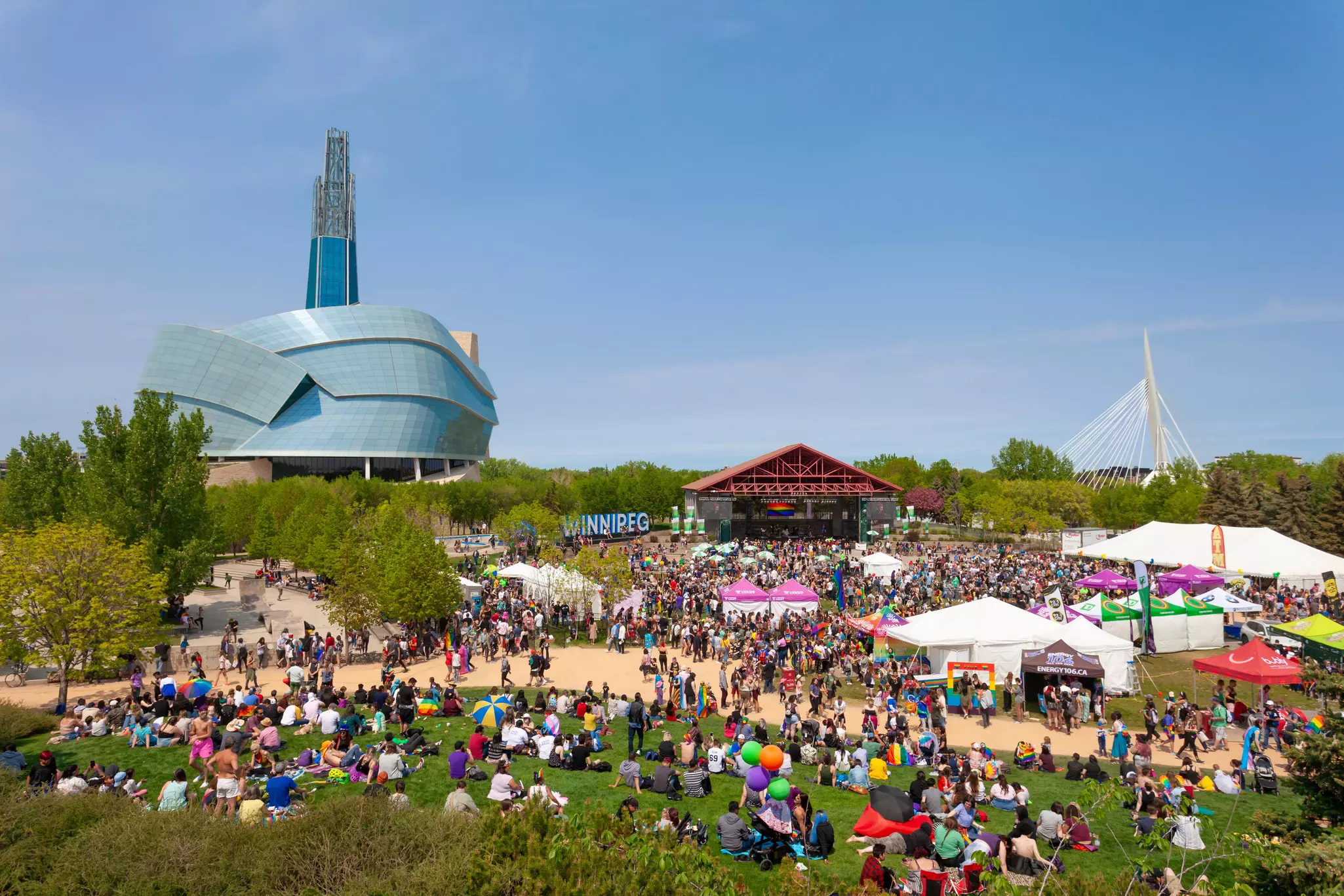A large assembly of people is gathered at the Forks to celebrate Pride Weekend 2019 in Winnipeg. The Canadian Museum for Human Rights can be seen in the back.