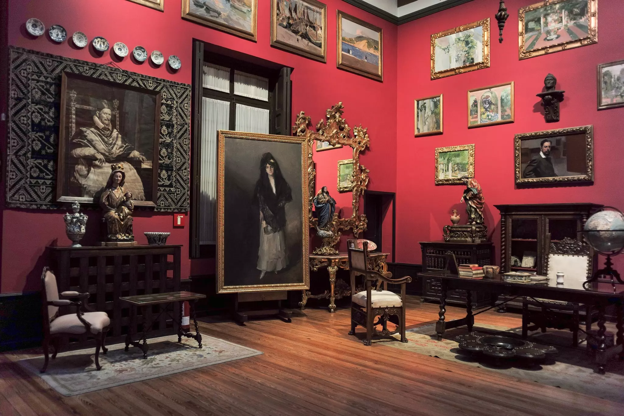 Paintings, chairs and other furnishings are displayed in a large gallery with red walls.