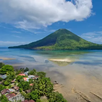 Green conical volcano across a turquoise channel, seen above a village of tin roofs and palm trees under a big white cloud.