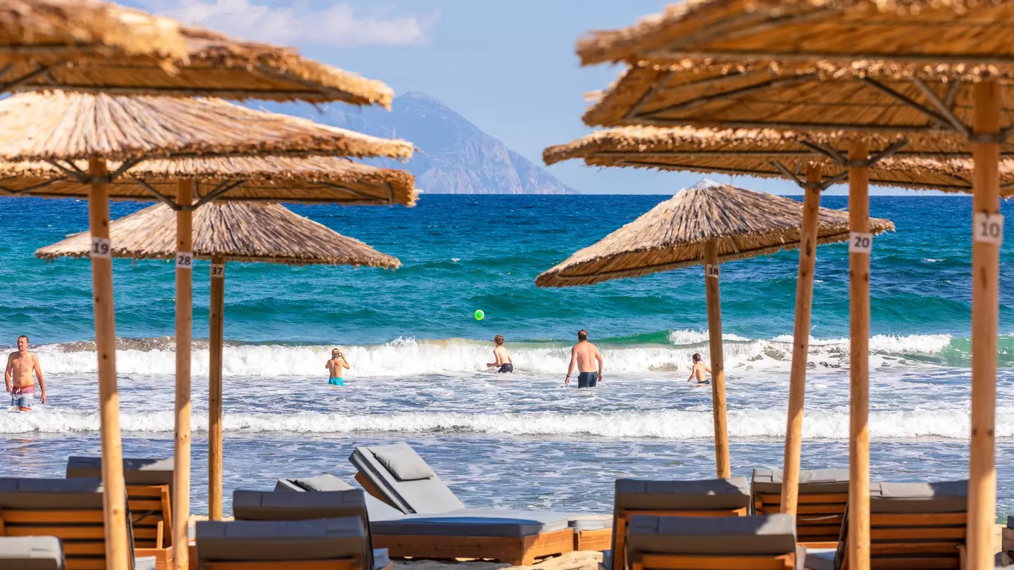 People splash about in the sea and play ball games. In the foreground are sun loungers and bamboo parasols.