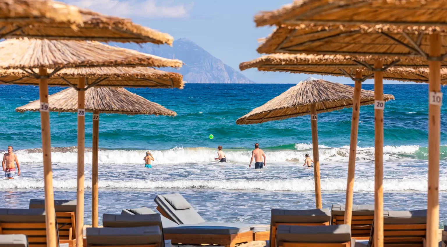 People splash about in the sea and play ball games. In the foreground are sun loungers and bamboo parasols.