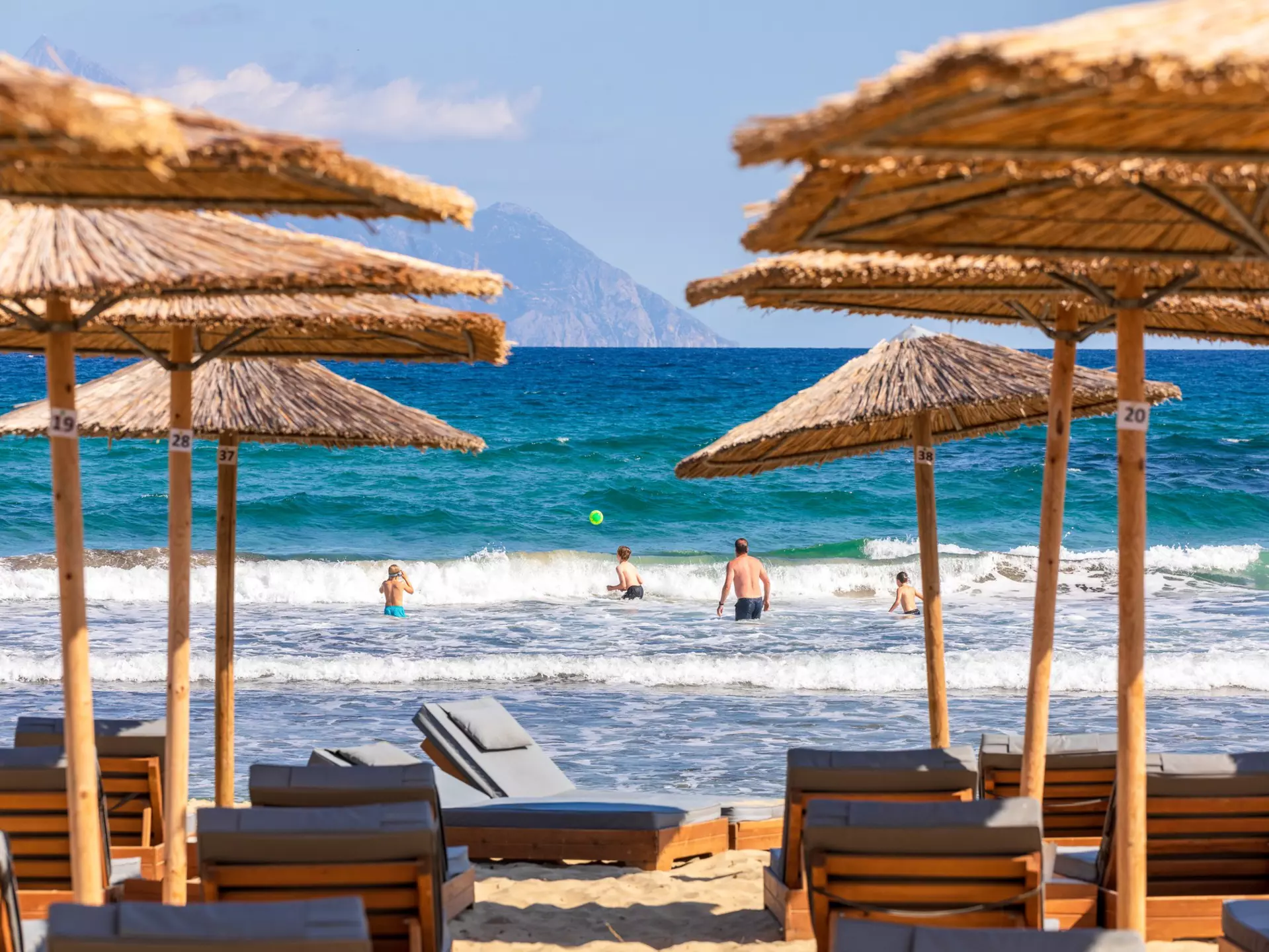 People splash about in the sea and play ball games. In the foreground are sun loungers and bamboo parasols.