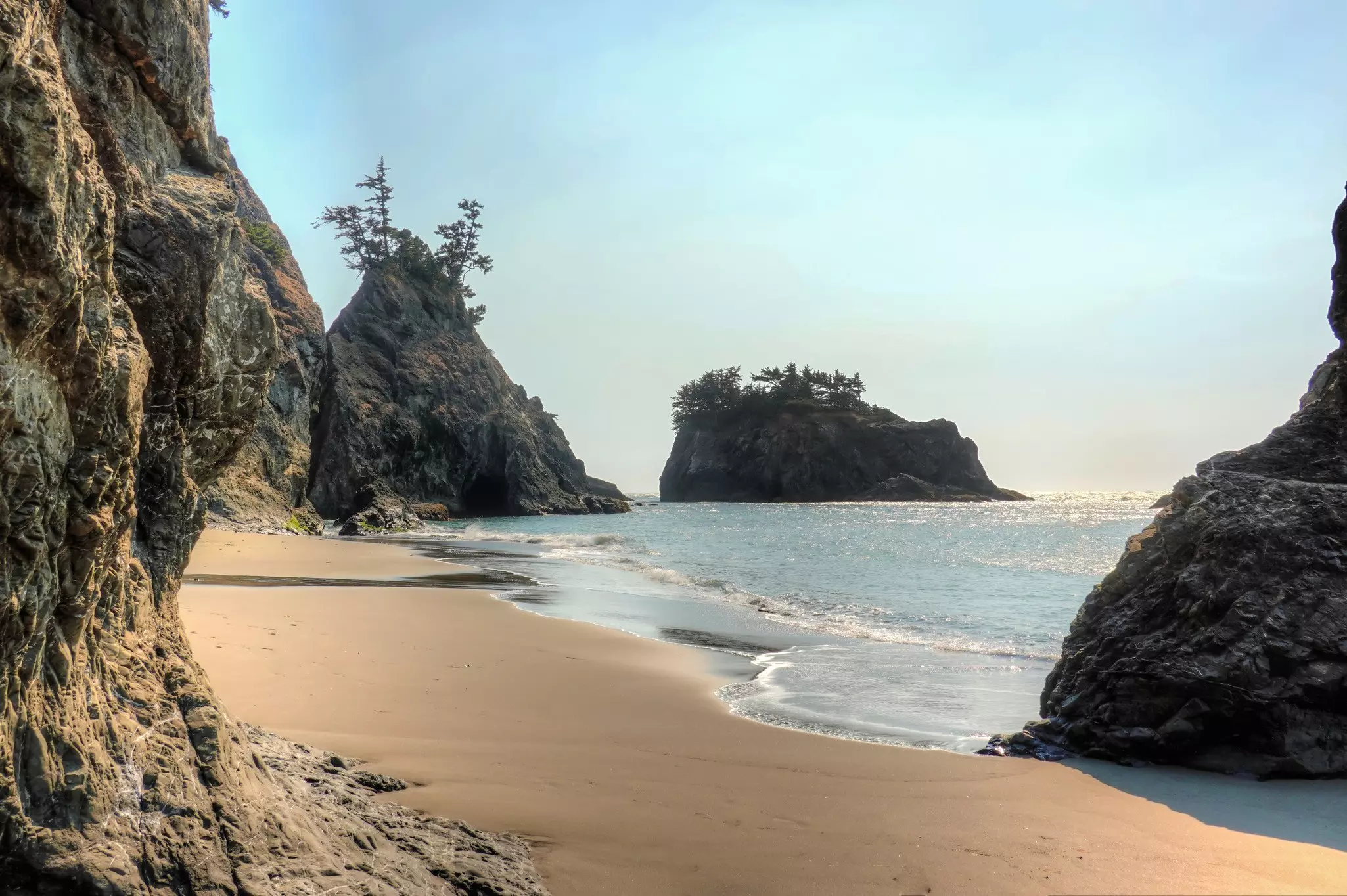 Tree-topped sea stacks near a sandy beach lapped by waves.