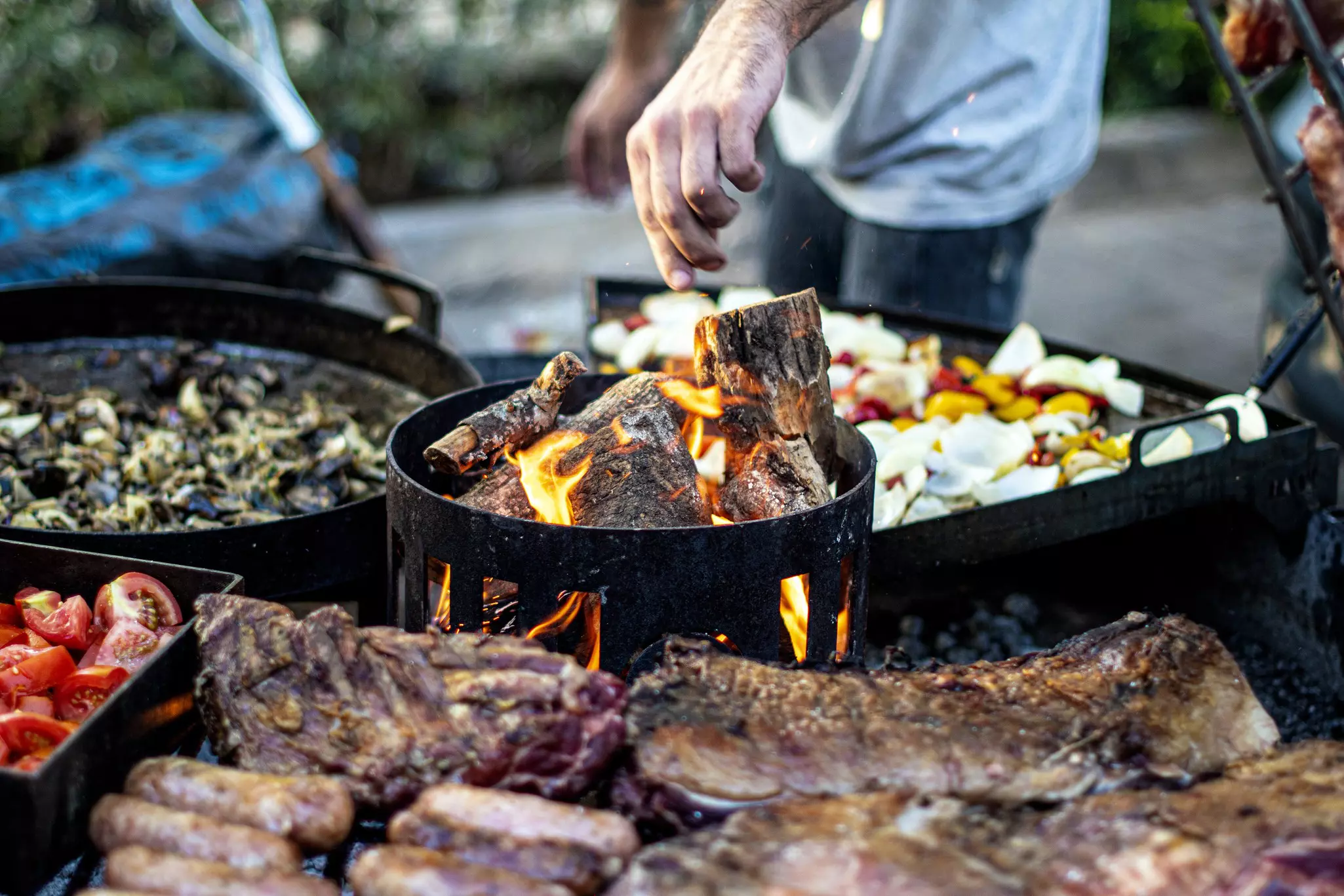A man is tending to a large, wood-fired barbecue grill is loaded with various meats and vegetables