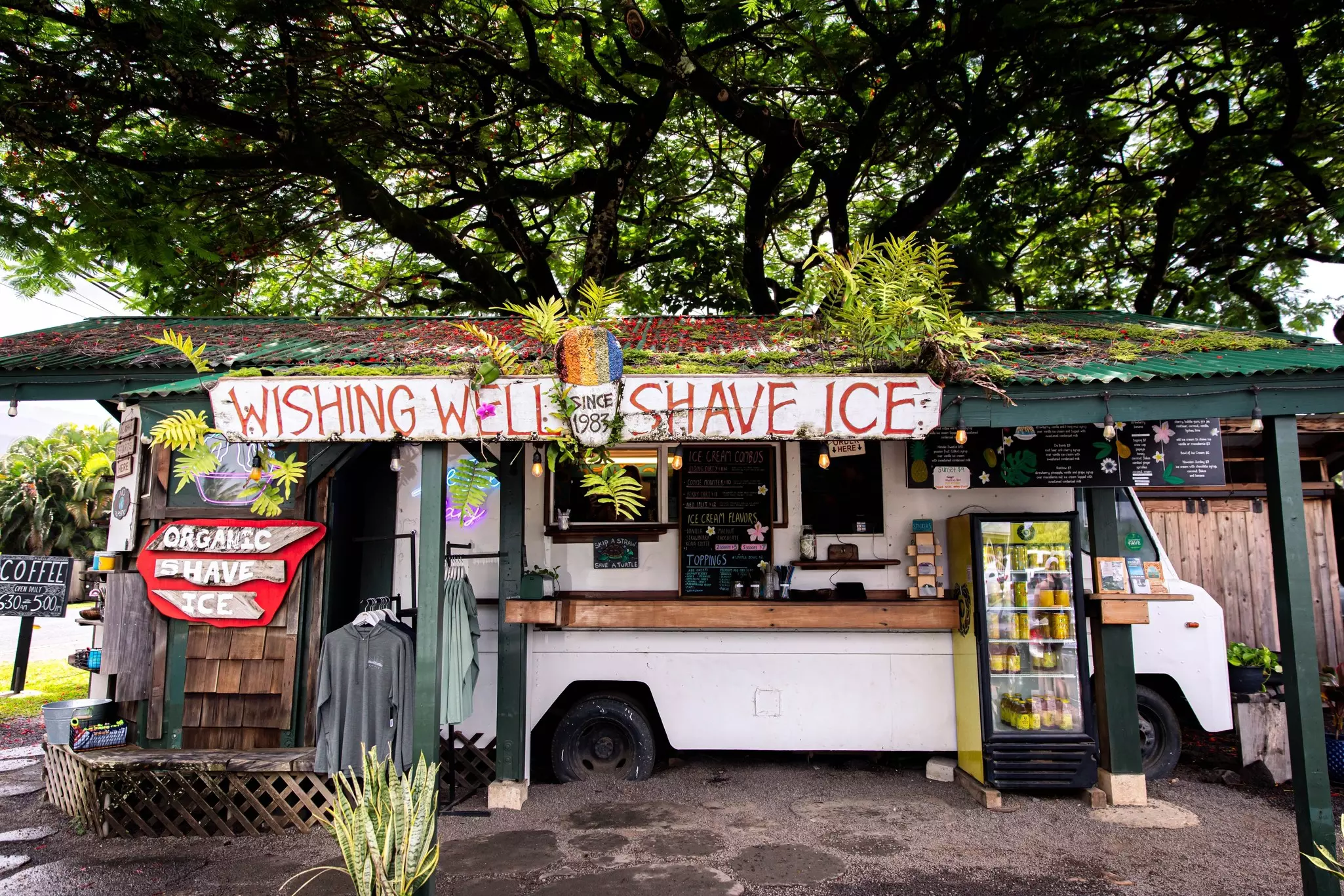 A white food truck backed under a shingled roof with shingled store to the left selling shave ice.