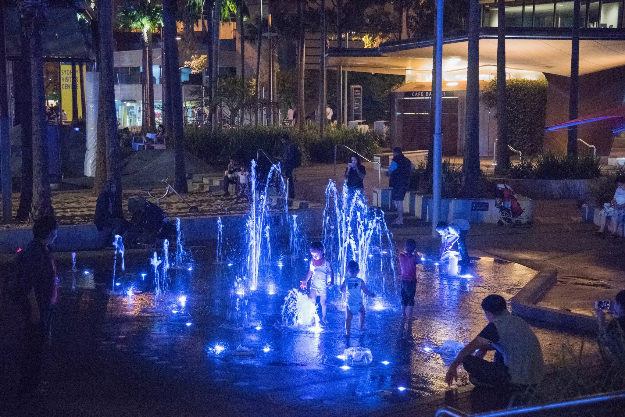 Children play at night in a water fountain illuminated with blue lights in a city.