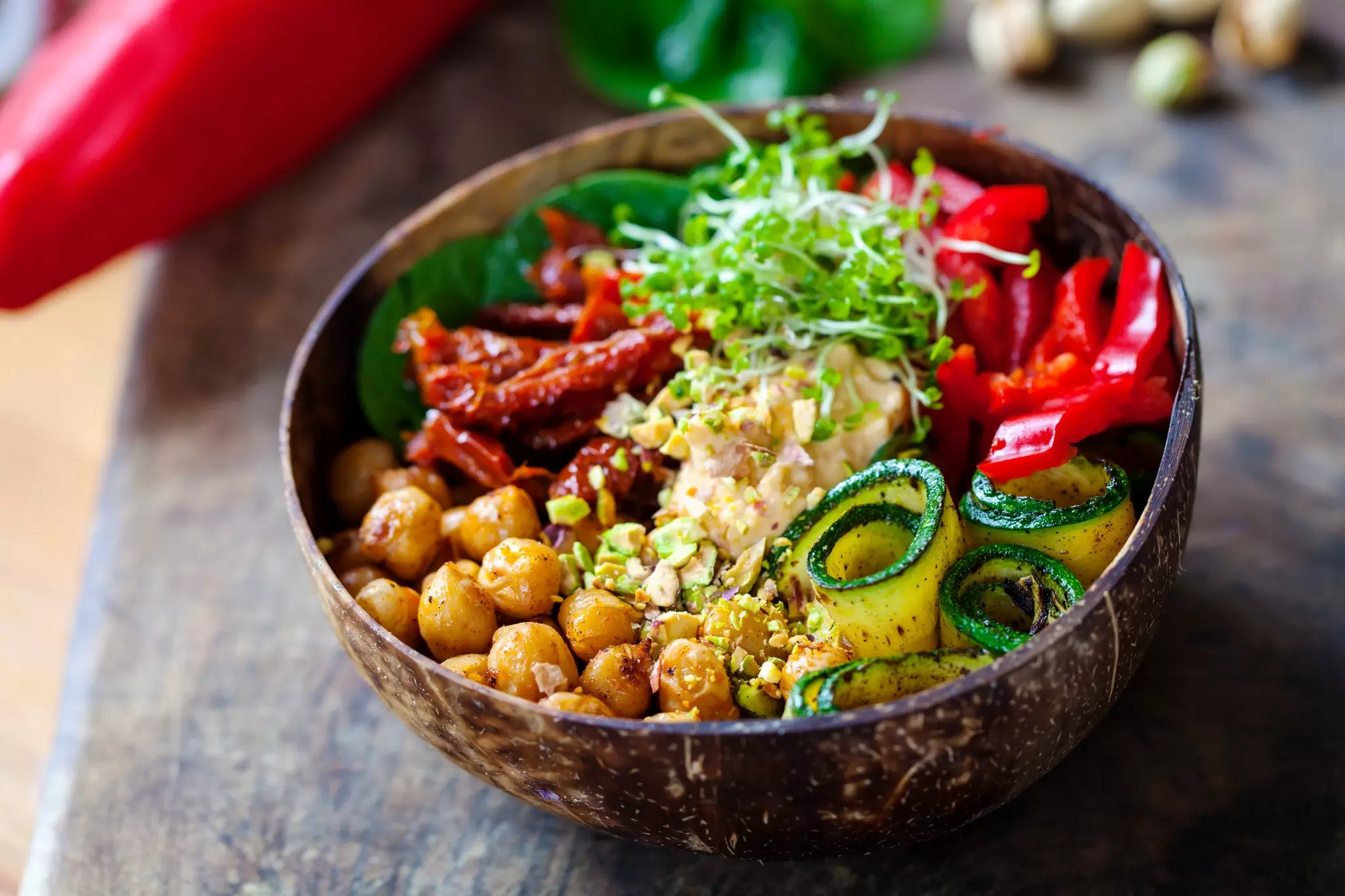 Coconut bowl filled with roasted chickpeas, hummus, rolled grilled zucchini, red pepper, sun-dried tomatoes and fresh sprouts.