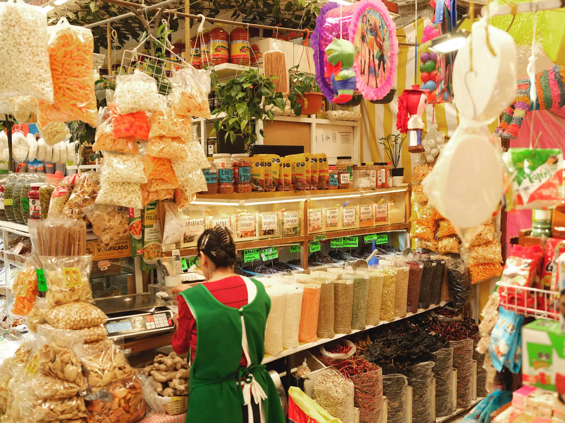 A vendor in a food market stands at a scale, surrounded by food and decor products like pinatas