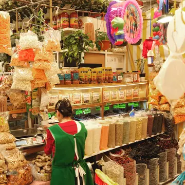 A vendor in a food market stands at a scale, surrounded by food and decor products like pinatas