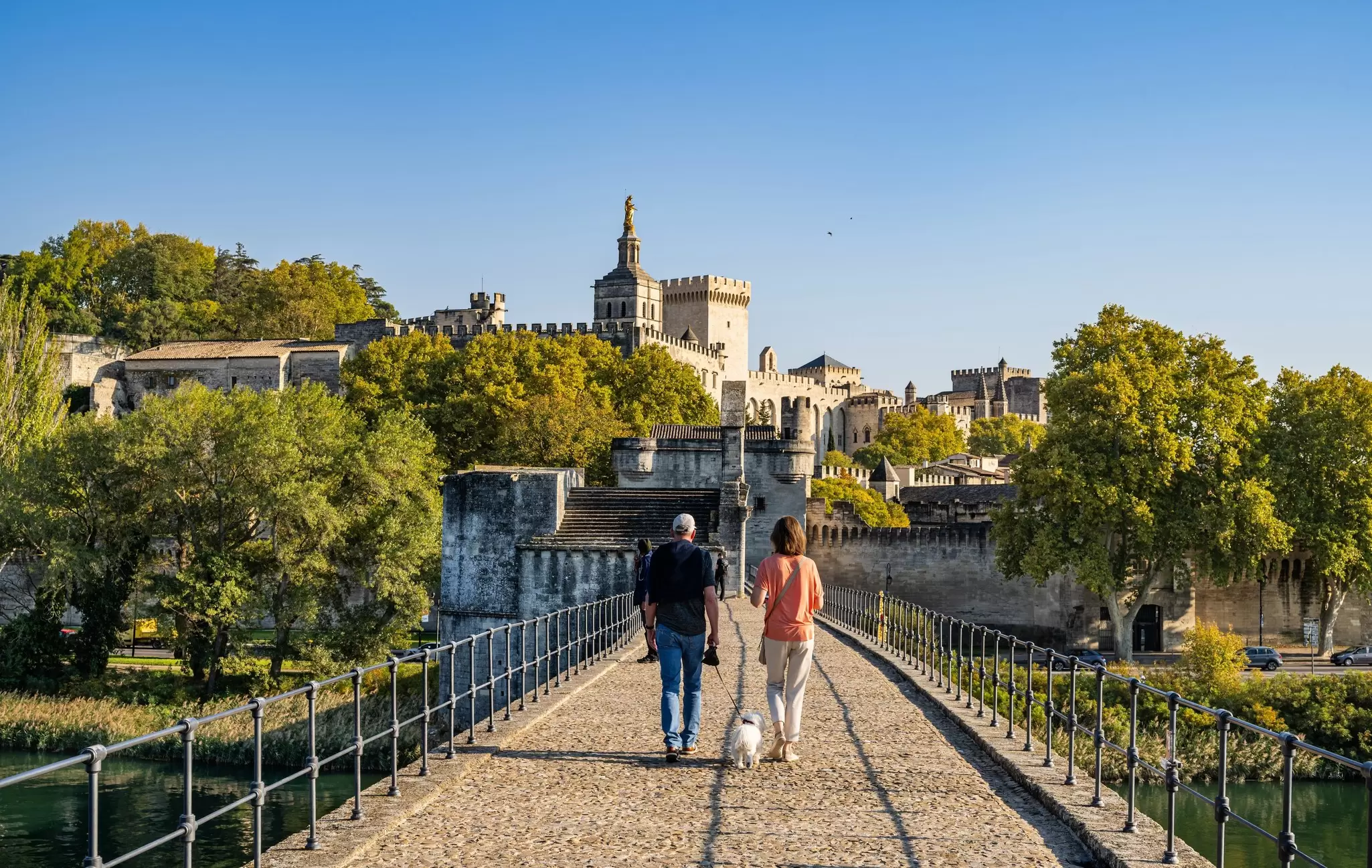 Two people walk on a bridge with a white dog toward a stone castle.
