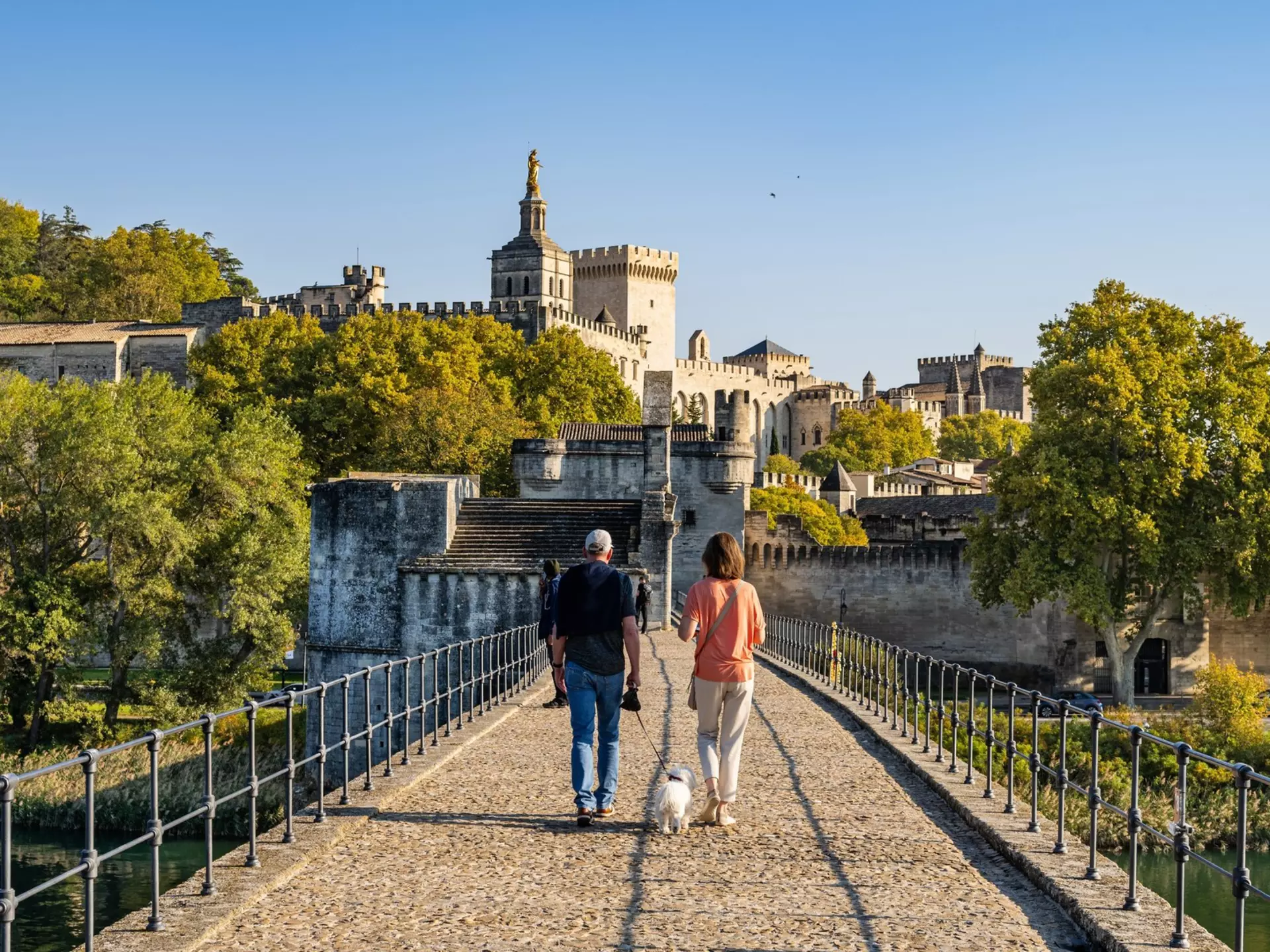 Two people walk on a bridge with a white dog toward a stone castle.