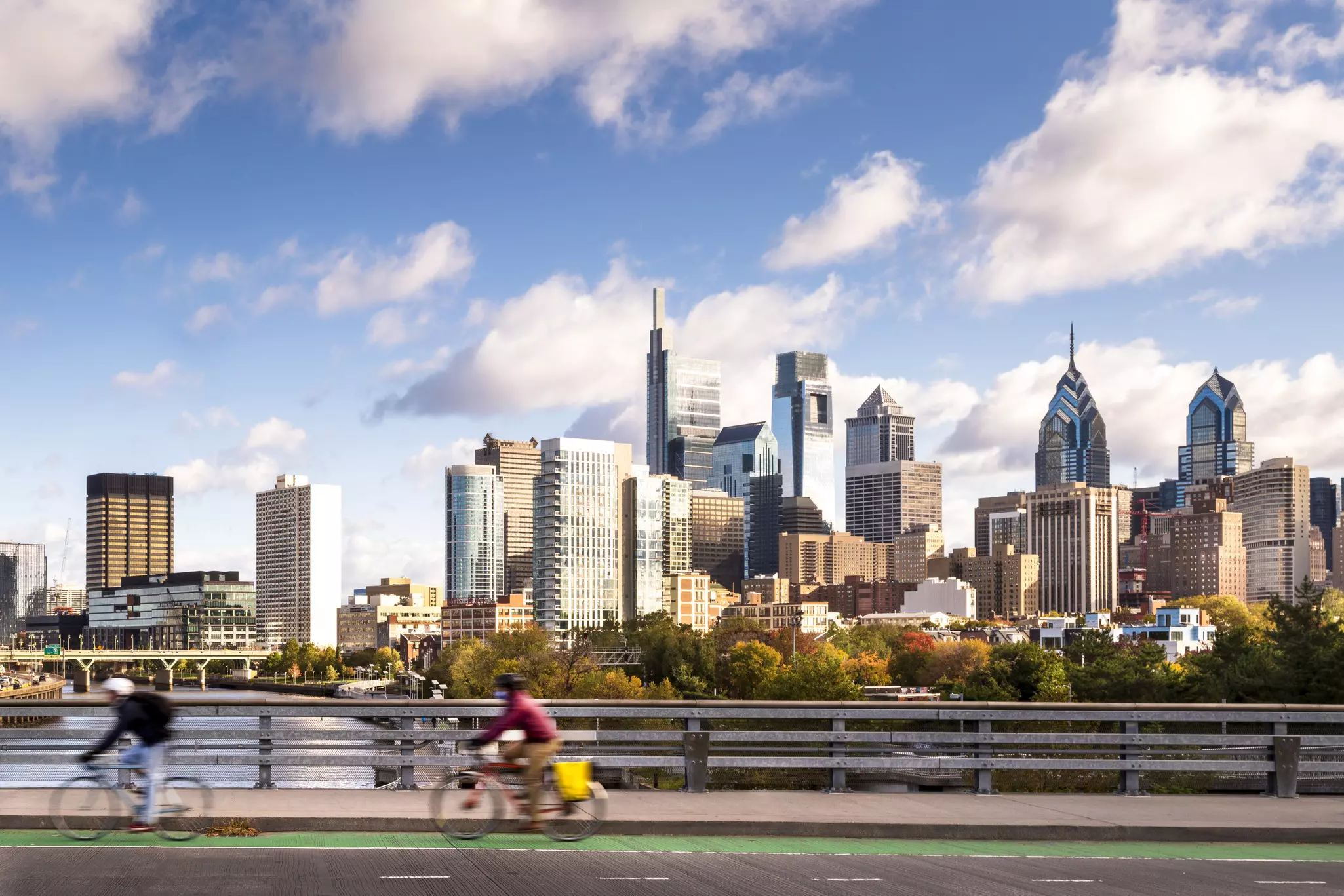 Two out-of-focus cyclists on a bridge with city skyline in the distance on a mostly sunny day.
