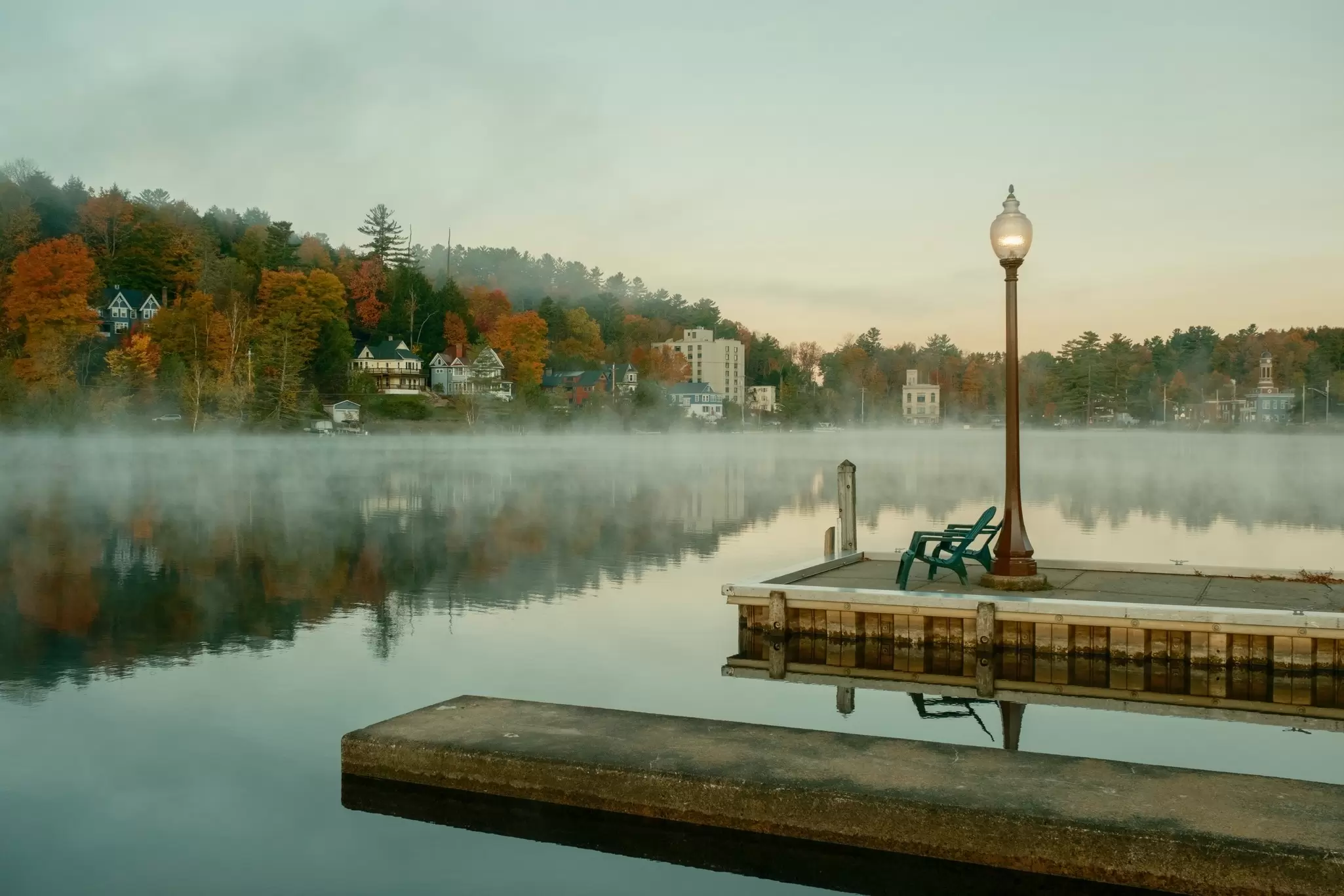 Morning fog over a lake; trees in autumn color are on the shore, and Adirondack chairs are on a dock.