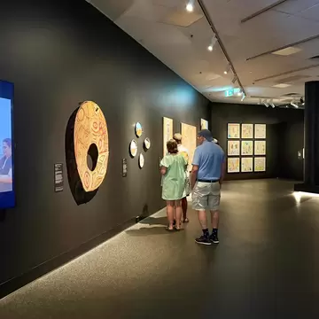 Visitors look at painted Aboriginal artworks on dark gallery walls, beside a video screen showing two people on a beach.