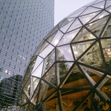 Partial view of a spherical glass structure with plants inside, in front of a tall building mostly made of glass