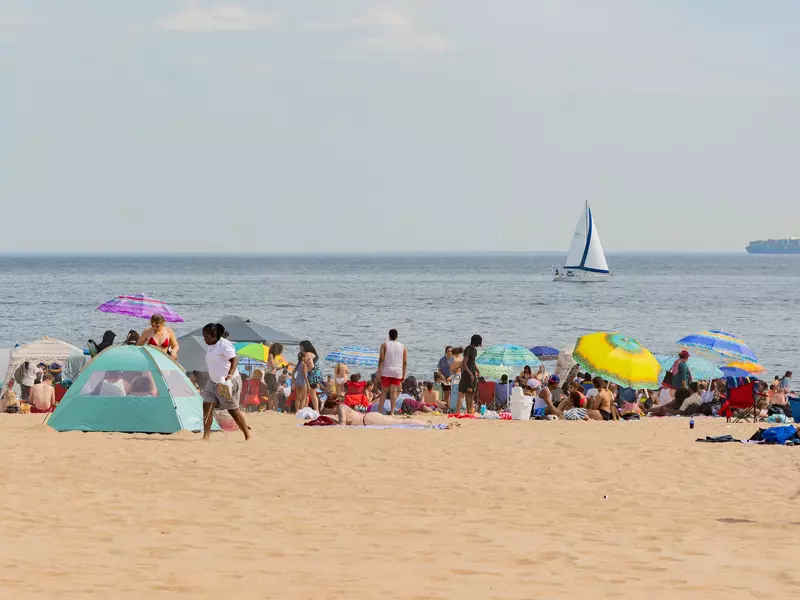 People with colorful umbrellas and sun tents on a sandy beach; a sailboat is in the water.