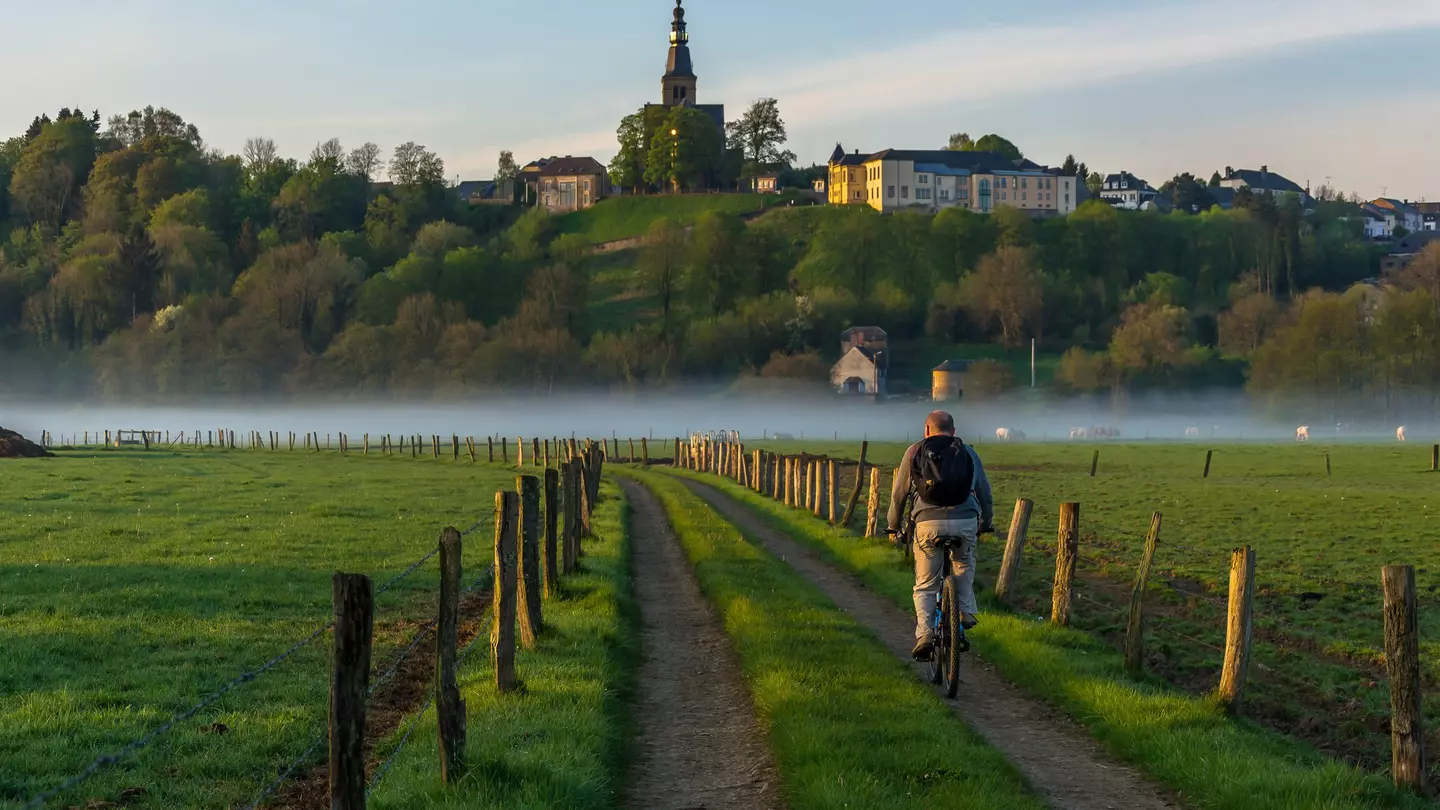 A man on a bicycle is pictured from behind as he pedals down a rural road past fields and toward a misty river. A church and houses are seen on a ridge in the distance.