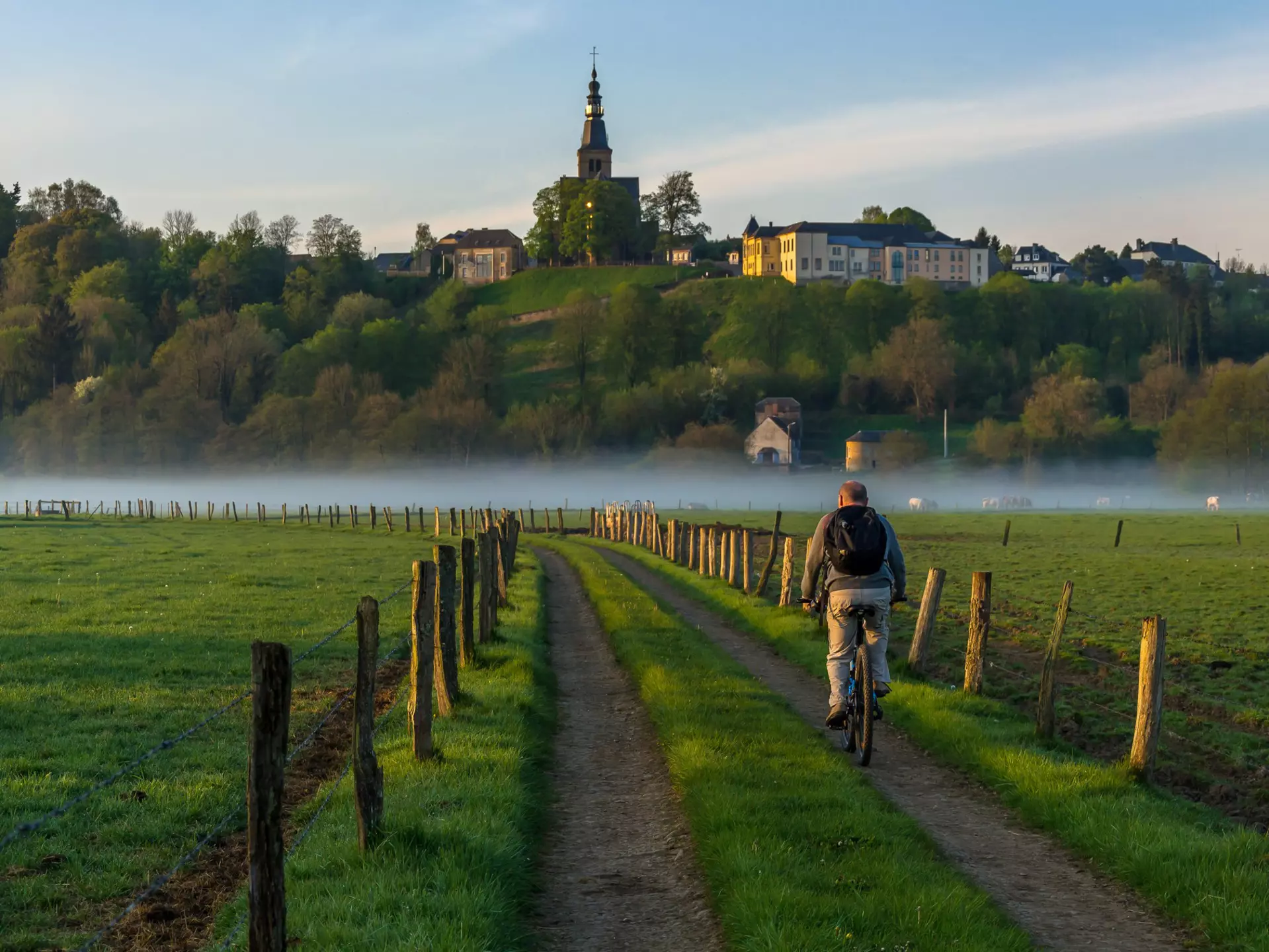 A man on a bicycle is pictured from behind as he pedals down a rural road past fields and toward a misty river. A church and houses are seen on a ridge in the distance.