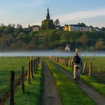 A man on a bicycle is pictured from behind as he pedals down a rural road past fields and toward a misty river. A church and houses are seen on a ridge in the distance.