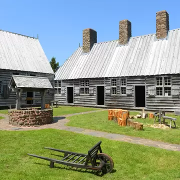 Grey wooden houses with stone chimneys around a grassy courtyard, a covered well and an old wooden wheelbarrow