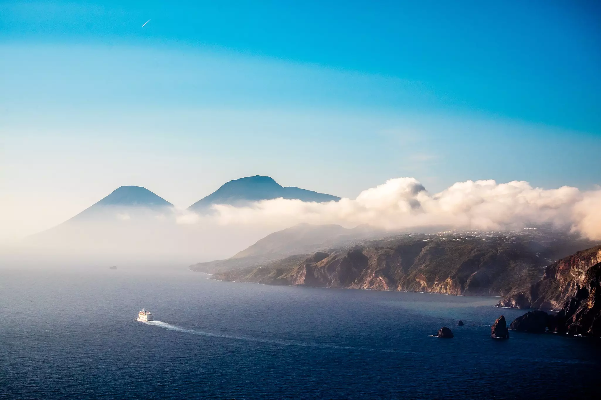 A ferry curves through the ocean between two volcanic islands with clouds rolling off the land and facing towards the ocean.