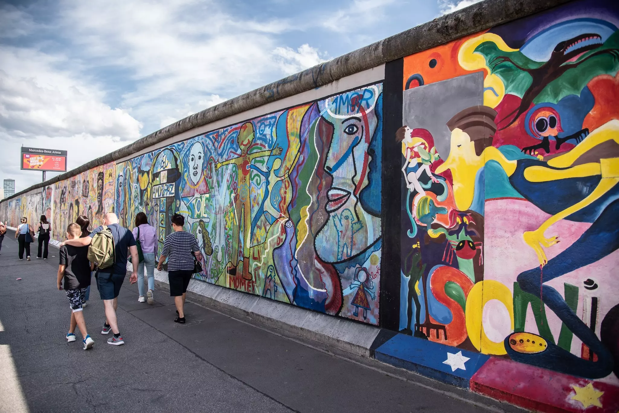 People walk on a concrete path alongside a concrete wall with colorful street art on a mostly sunny day.