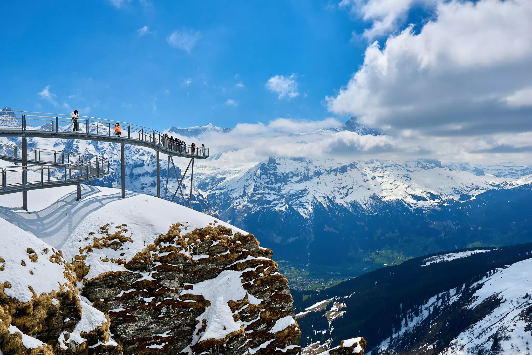 People on a glass walkway installed on a cliff overlooking snow-covered mountains